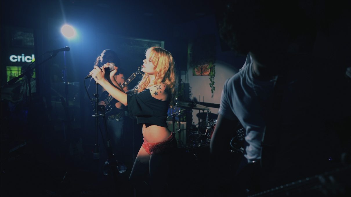 A shop-floor set lit almost entirely blue, the singer centre frame with both hands on the mic, guitarist and drummer behind her in shadow